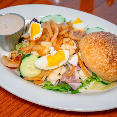 Chef salad with hard-boiled eggs, ham, cucumbers, dressing, and a sesame bun.