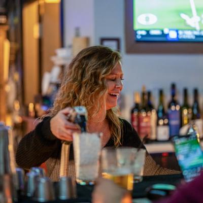 A bartender smiles while pouring a drink at a lively bar.