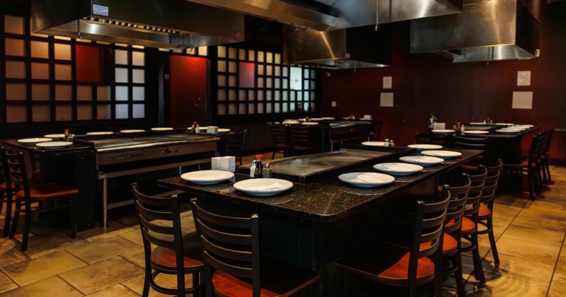 A teppanyaki restaurant with grill tables, white plates, black chairs and ventilation hoods.