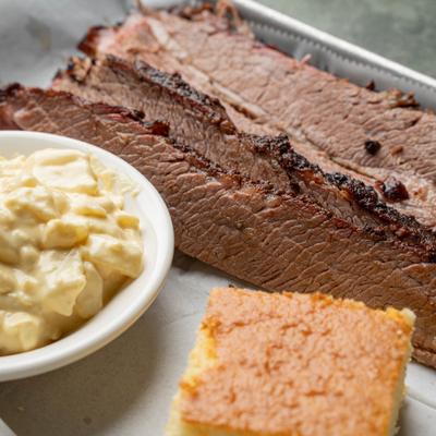 Sliced smoked beef brisket served with potato salad and a square of cornbread.