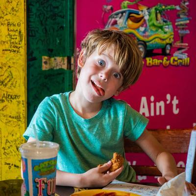 A kid sitting at a table with food, enjoying a meal and making a funny face for the camera.