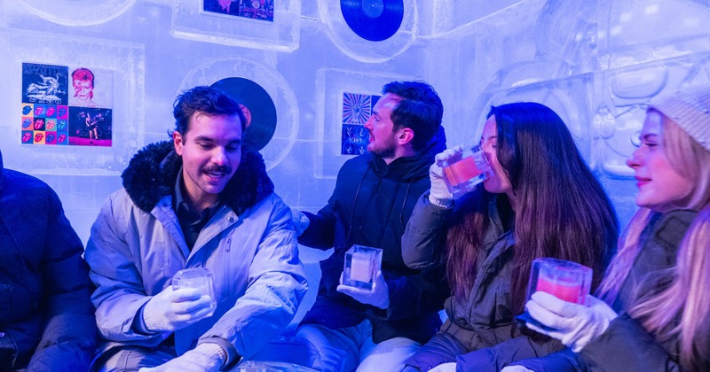 A group of people in winter attire enjoy drinks in an ice-themed bar