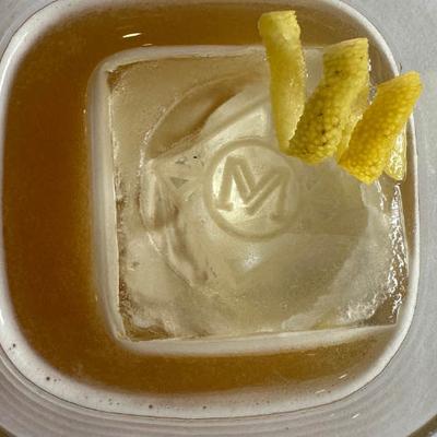 A cocktail drink garnished with Mere Blues logo engraved ice cube and lemon peel, top view.