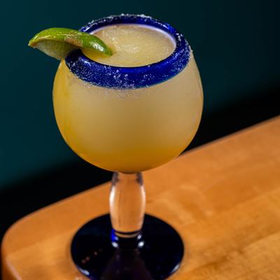 A frozen margarita  served in a stemmed glass with a blue-rimmed top and a lime wedge garnish.