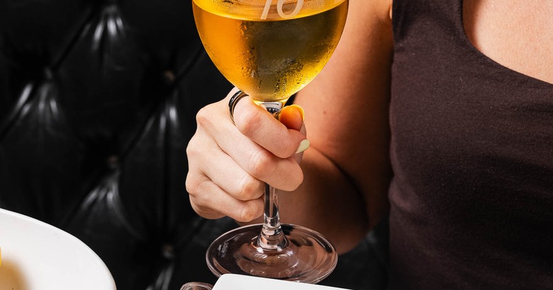 A person holding a glass of white wine at a table with a plate of food