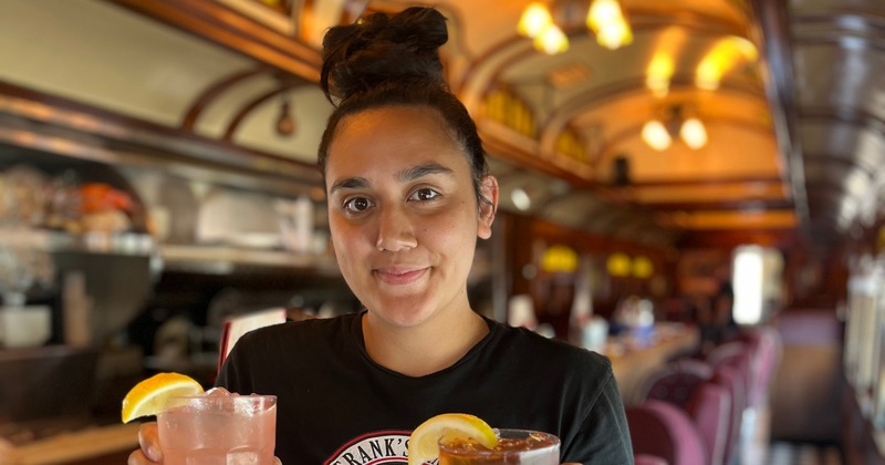 Smiling staffer holding two drinks with lemon wedges in a cozy, warmly lit diner