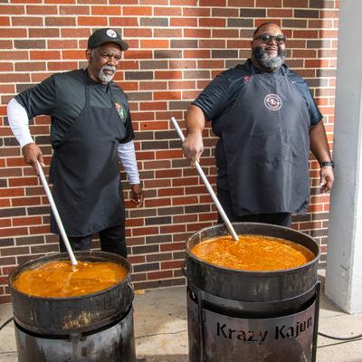 Two cooks stirring large pots of stew.