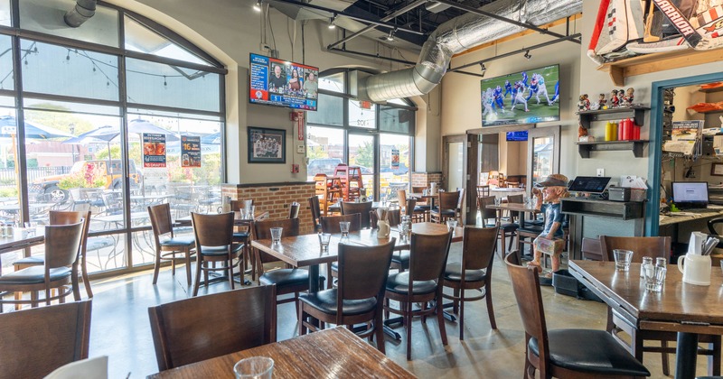 Interior of a bright restaurant with wooden tables and chairs, large windows, and mounted televisions