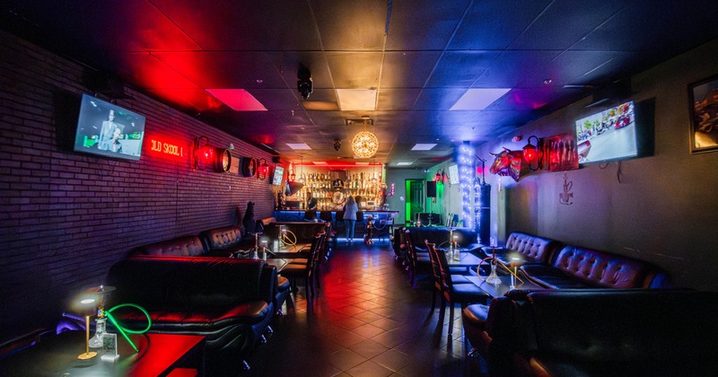 Dimly lit bar lounge with black leather couches, red and blue lighting, brick walls, and a bar