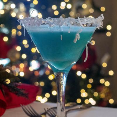 A light blue cocktail in a martini glass with a coconut-rimmed edge, festive background.