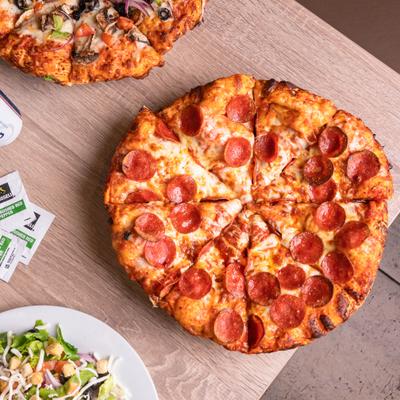 Pepperoni pizza on a wooden table beside a veggie pizza and a salad.