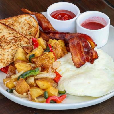 Breakfast plate featuring fried eggs, bacon, home fries, toasted bread, and two dipping sauces.