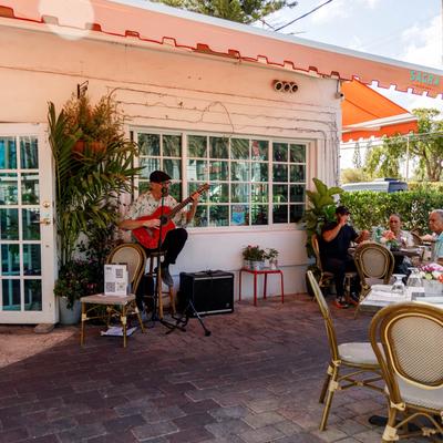 A musician plays guitar outside, Nearby, diners at a table enjoy meal.