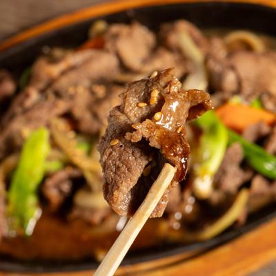 Piece of beef being picked up with a chopstick, above the skillet of Sizziling beef.
