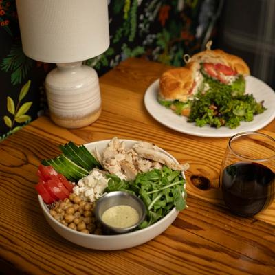 It's All Greek To Me grain bowl, a glass of red wine, and a plate with chicken salad crissant.