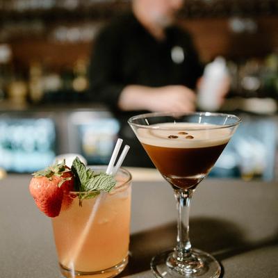 Two cocktails on a bar counter with a blurred bartender in the background.