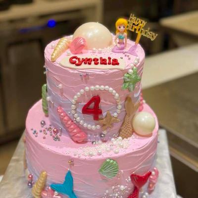Two-tiered pink mermaid birthday cake decorated with shells, pearls, and the name Cynthia.