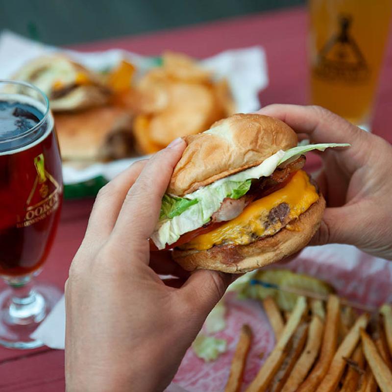 Person holding a Bacon Cheeseburger above the table with food and beer.