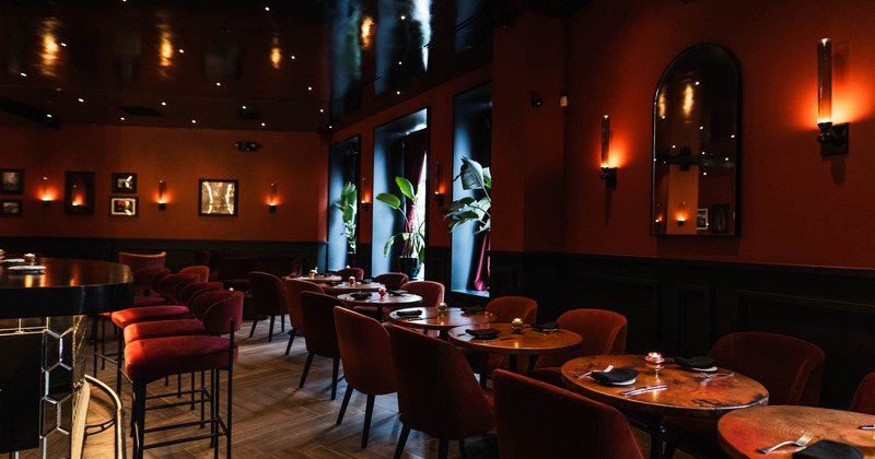 A dimly lit restaurant interior with red walls, round wooden tables, and plush red chairs