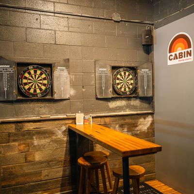 Dartboards and a wooden bar table with stools in a game room.