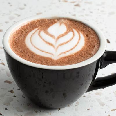 Black cup of cappuccino with leaf-shaped latte art sits on a speckled white countertop.