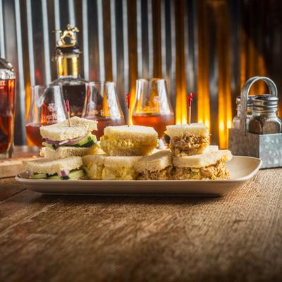 A platter of assorted small sandwiches served with amber drinks in rustic lighting.