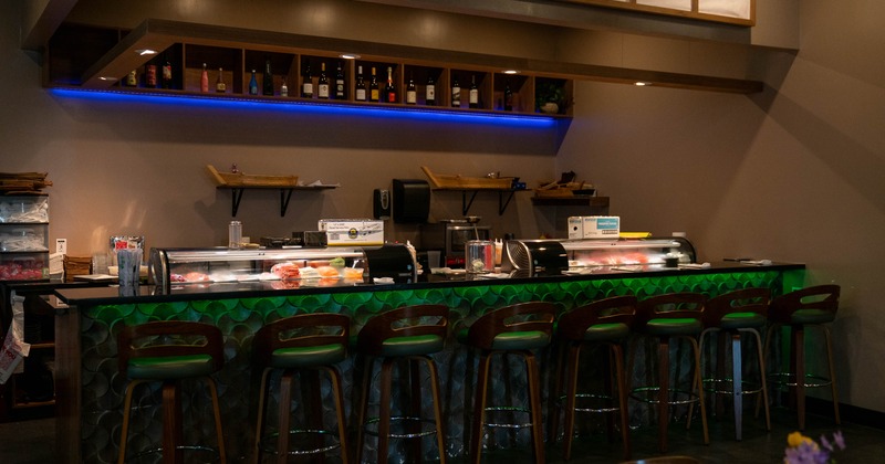 A dimly lit sushi bar - green-patterned counter, bottle shelves, wooden bar stools