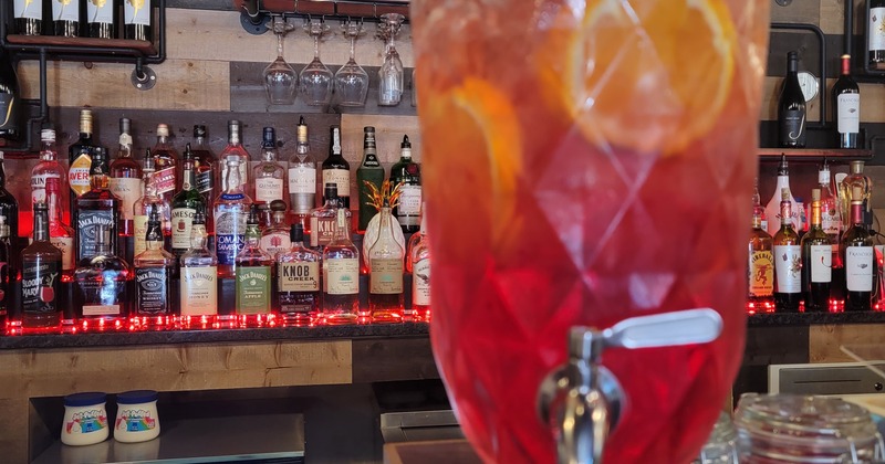 A red drink dispenser on a bar counter, with various bottles of liquor in the back bar