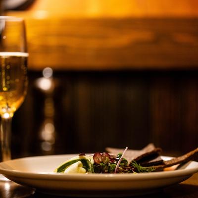 A glass of sparkling wine next to a plate of food garnished with herbs on a dimly-lit table.