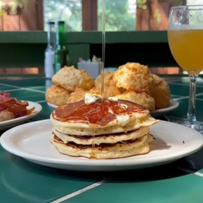 Pancakes with syrup served on green tiled table, accompanied with other dishes and a drink.