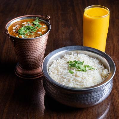 Traditional lentil dish served in a copper pail, next to a silver bowl of rice and Mango Lassi.