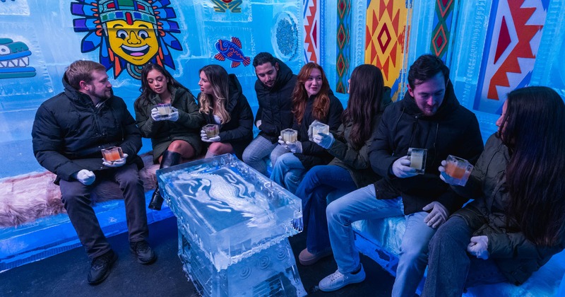 A group of guests holding drinks around a carved ice table inside a brightly lit blue ice bar