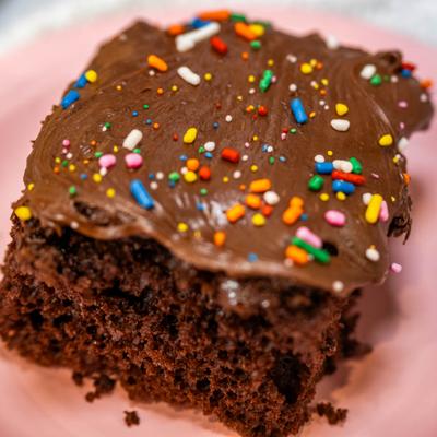 Sllice of chocolate cake topped with chocolate icing and rainbow sprinkles.