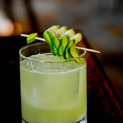 A refreshing pale green cocktail, garnished with a curled cucumber slice on a toothpick.