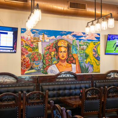 Dining room , banquette seating and tables by a wall with TVs and a large painting of Frida Kahlo.