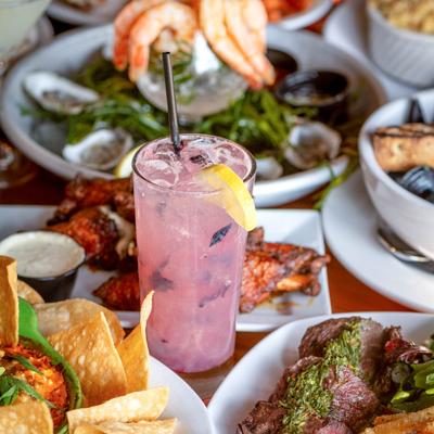 A glass of Unity of Maine cocktail sits on a wooden table, surrounded by food plates.