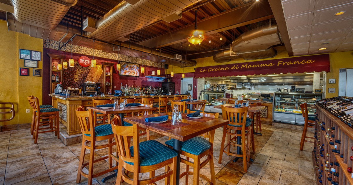 Interior with wooden tables, blue cushioned chairs, a bar and a visible kitchen.