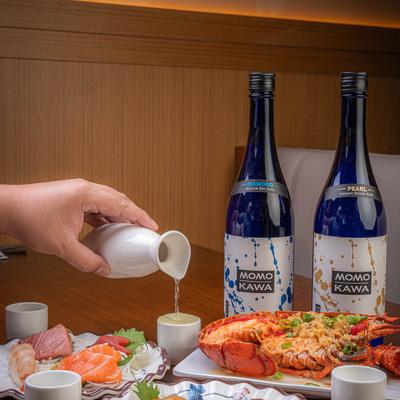 A person pours sake into a cup on a table with multiple dishes and sake bottles.