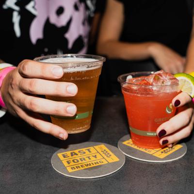 Two people hold a plastic cup of beer and a red-orange iced drink..