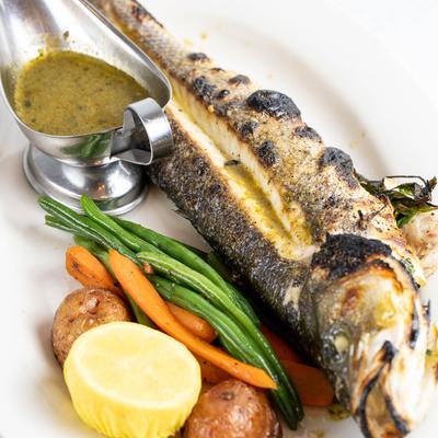 Grilled branzino with veggies, lemon half and sauce in a a metal gravy boat.