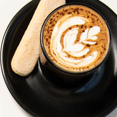 Cappuccino with latte art served in a black cup with a ladyfinger biscuit.