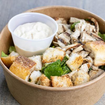 Grilled chicken Caesar salad with a cup of sauce in a paper bowl.