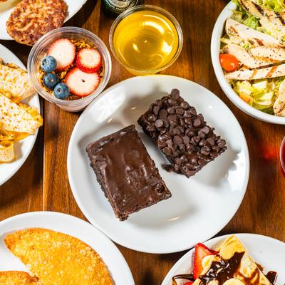 Two chocolate brownies on a plate surrounded by assorted dishes and drinks.