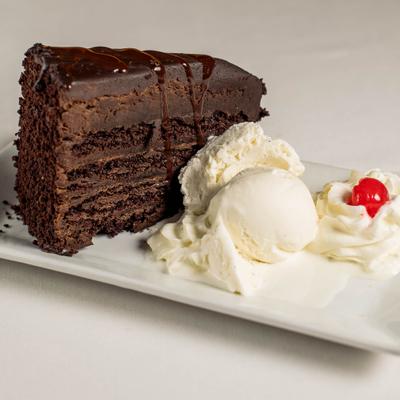 Rich chocolate layer cake slice plated with vanilla ice cream, whipped cream and a cherry.