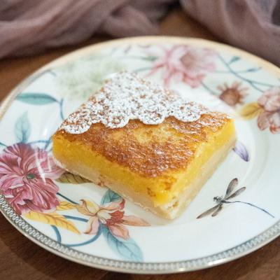 A square lemon bar with powdered sugar is on a floral plate.