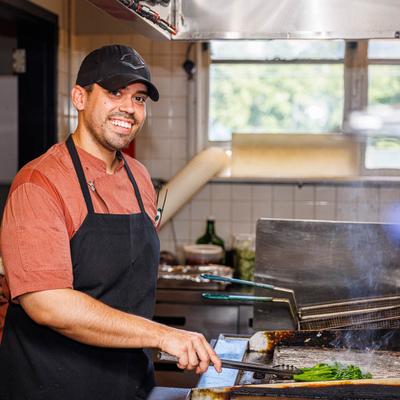 A smiling chef in a black apron and cap grills vegetables on a stove in a bright kitchen.