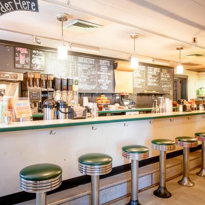 Retro diner counter with green vinyl stools, hanging pendant lights, chalk menu boards.