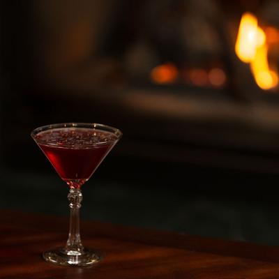 Red cocktail served in a martini glass by a fireplace.