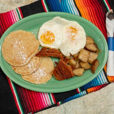 A green plate holds pancakes, fried eggs, bacon, and crispy breakfast potatoes.