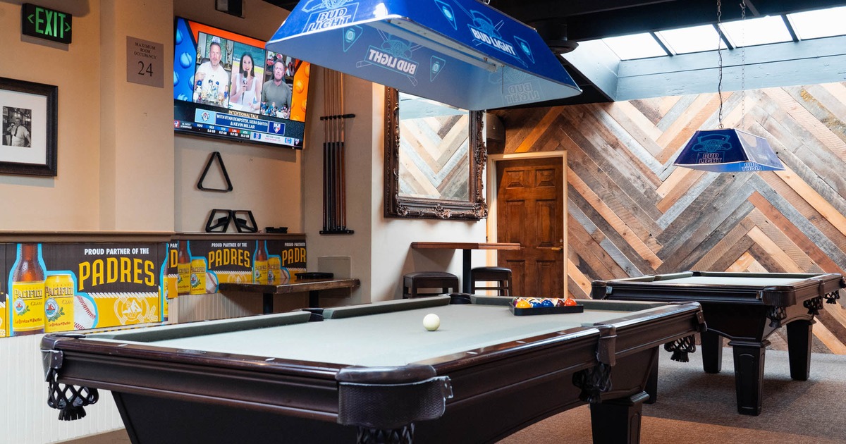 Interior, two pool tables, a wall-mounted TV showing sports, cue rack
